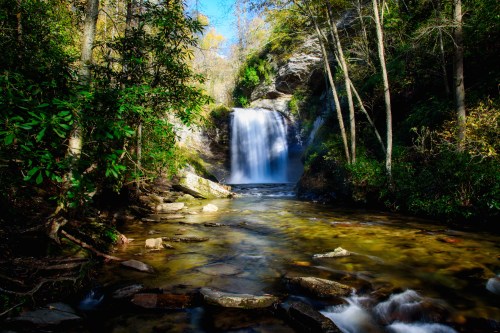 A serene waterfall cascades into a clear stream, surrounded by lush greenery and rocky terrain under a bright blue sky.