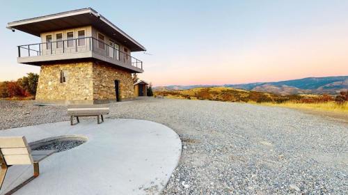 A stone building with large windows sits on a gravel area, overlooking rolling hills at sunset.