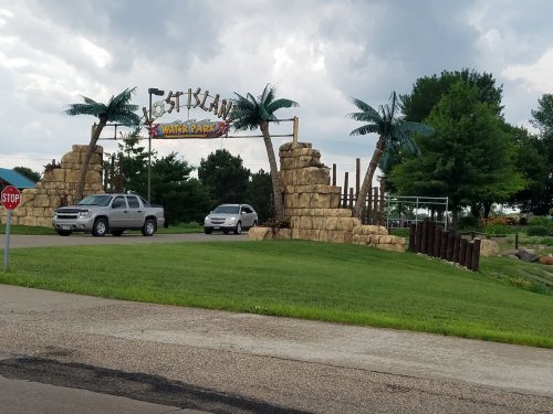 Entrance to a water park with palm trees and a sign reading "Fish Island Water Park," surrounded by green grass.