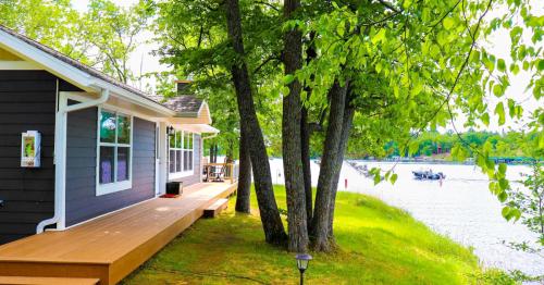 A serene lakeside view featuring a house, deck, trees, and boats on the water under a clear blue sky.