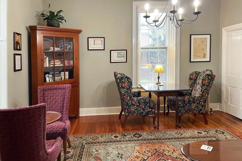 Cozy room with floral chairs, a wooden table, a bookshelf, and a chandelier, featuring warm lighting and a rug.