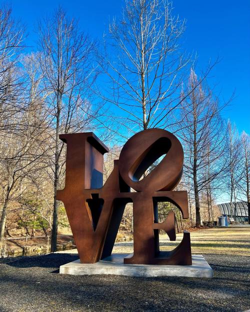 A large, rust-colored sculpture of the word "LOVE" stands in a park with bare trees and a clear blue sky.