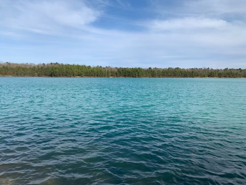 A tranquil lake with clear turquoise water, surrounded by lush green trees under a partly cloudy sky.