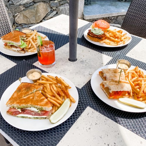 Four plates of sandwiches and fries on a table, with a drink in a glass, set outdoors.