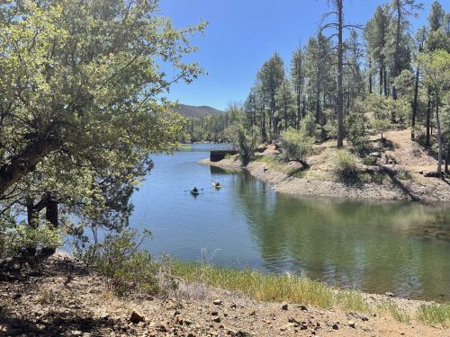 A serene lake surrounded by trees, with two kayakers paddling on the calm water under a clear blue sky.