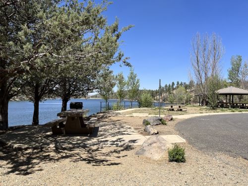 A serene lakeside park with picnic tables, trees, and a clear blue sky.