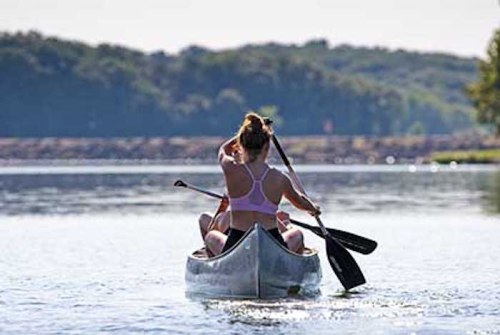 A person paddles a canoe on a calm lake, surrounded by trees and a clear sky.