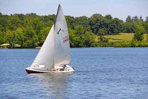 A small sailboat glides across a calm lake, surrounded by lush green trees and a clear blue sky.