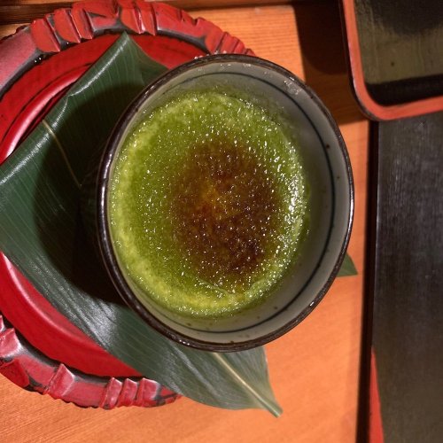 A bowl of green matcha dessert with a caramelized top, placed on a decorative red plate with a leaf underneath.