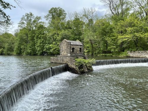 A stone building sits on a small island in a river, with a waterfall cascading over a dam surrounded by lush greenery.