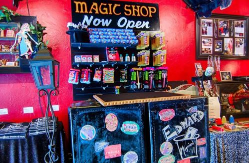 A vibrant magic shop interior with a "Now Open" sign, shelves of magic supplies, and a black counter.