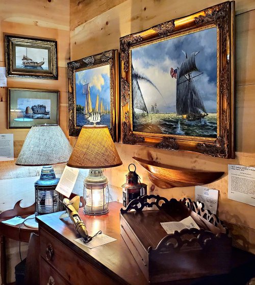 A cozy room with wooden walls, featuring nautical paintings, lamps, and a vintage desk with decorative items.