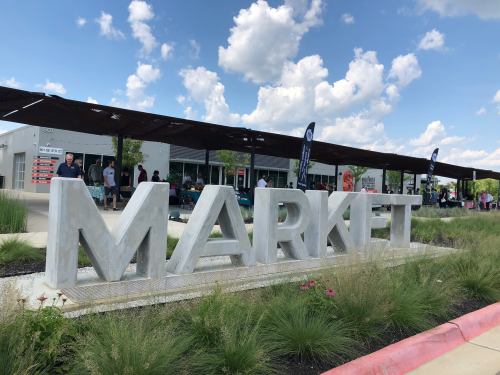Large concrete letters spelling "MARKET" in front of a modern building with people and greenery in the background.