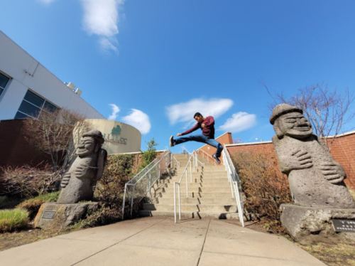 A person leaps over stairs flanked by two large stone sculptures under a blue sky with scattered clouds.