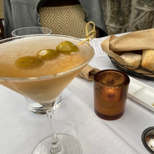 A close-up of a cocktail with olives, a candle, and a basket of bread on a dining table.