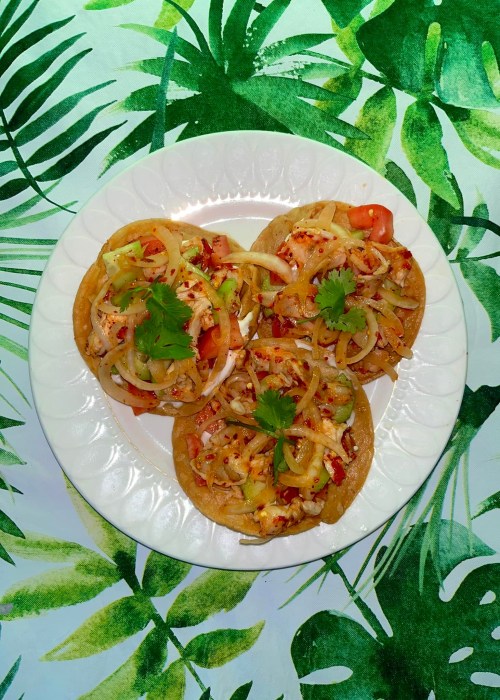 Three colorful tostadas topped with vegetables and cheese, served on a white plate against a green leafy background.