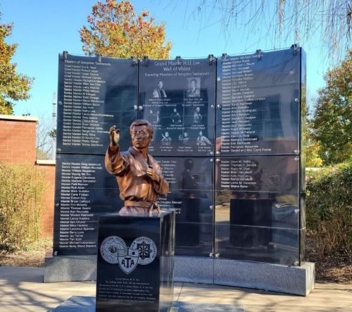 A bronze statue of a martial artist stands in front of a wall listing martial arts achievements and honorees.