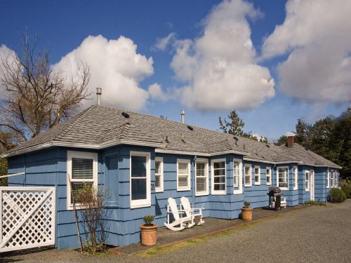 A blue cottage with a gravel driveway, featuring white chairs and potted plants under a partly cloudy sky.