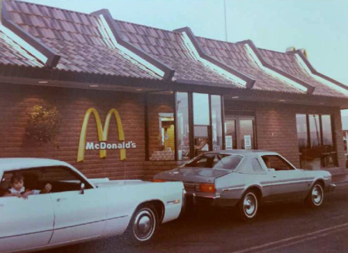A vintage McDonald's with two cars parked outside, one white and one gray, in a retro setting.