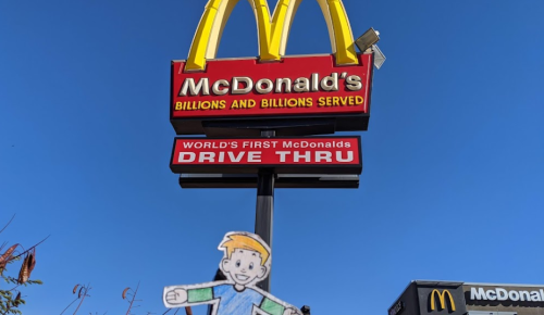 A colorful McDonald's sign against a blue sky, featuring a cartoon character in front of the world's first drive-thru.