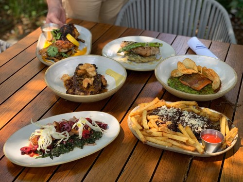 A table with five plates of diverse dishes, including grilled meats, vegetables, and fries, set outdoors.