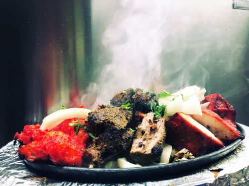 Sizzling platter of grilled meats, onions, and vibrant red sauce, with steam rising against a metallic background.