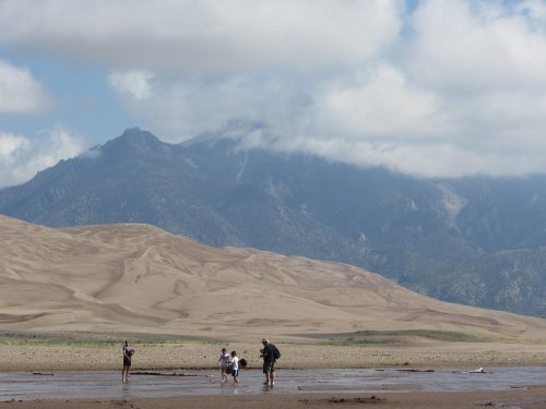A group of people wading in a shallow stream with sand dunes and mountains in the background under a cloudy sky.