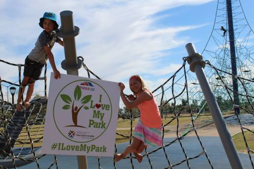 Two children play on a climbing structure at a park, holding a sign that says "I Love My Park."
