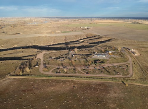 Aerial view of a rural area with a winding road, scattered buildings, and fields in the background under a clear sky.