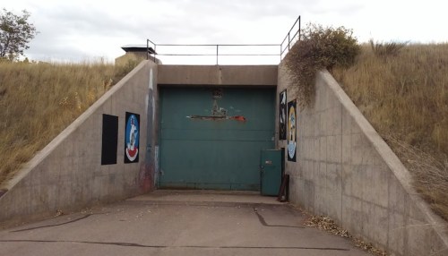 A large green door set into a concrete structure, flanked by murals, with a grassy hillside in the background.