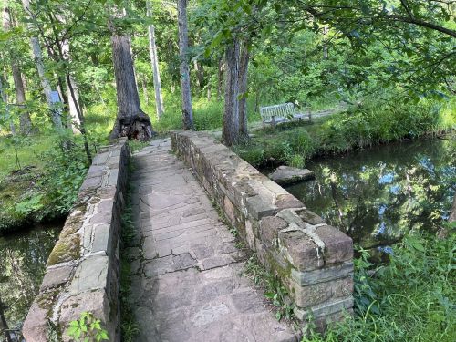 A stone bridge over a small stream, surrounded by lush greenery and trees, with a bench visible in the background.