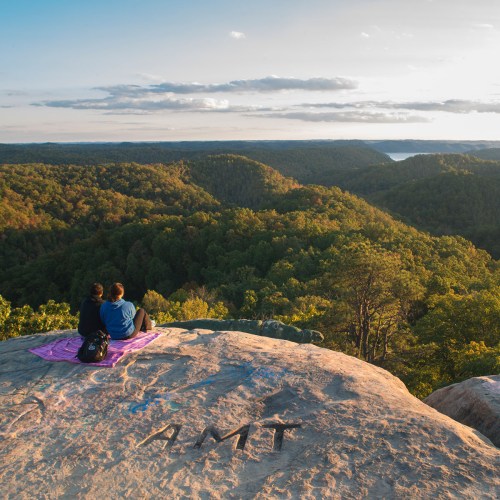 Two people sit on a rock overlooking a lush green landscape and a distant body of water during sunset.