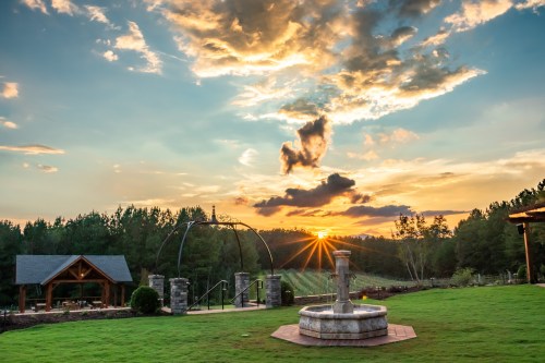 A scenic view of a sunset over a grassy area with a fountain, surrounded by trees and a rustic pavilion.
