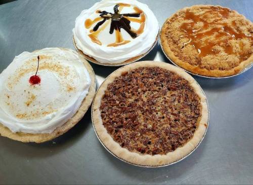 Four delicious pies on a silver surface: a cream pie with a cherry, a caramel pie, a crumb pie, and a pecan pie.