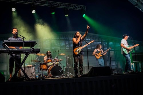 A live band performs on stage with a keyboard, guitars, and drums, under colorful stage lights.