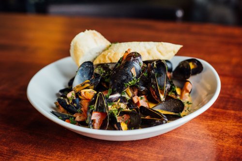 A bowl of mussels with herbs and diced tomatoes, served with a slice of bread on a wooden table.