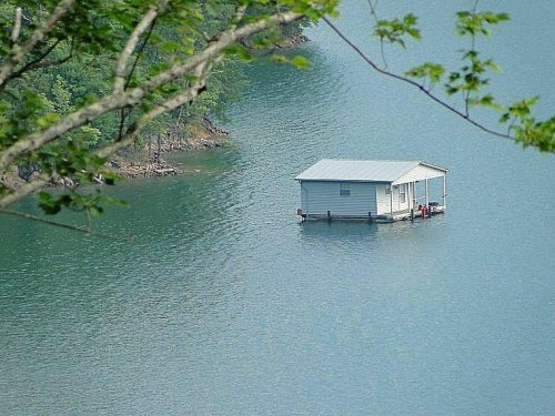 A small, white houseboat floats on calm water, surrounded by lush green trees and a serene landscape.