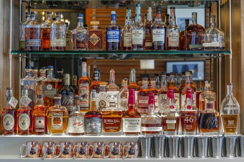 A display of various whiskey bottles on shelves, with copper mugs in front.