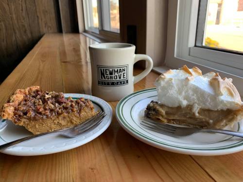 A wooden table with two plates of pie: one pecan pie and one cream pie, alongside a coffee mug from Newman Grove City Café.