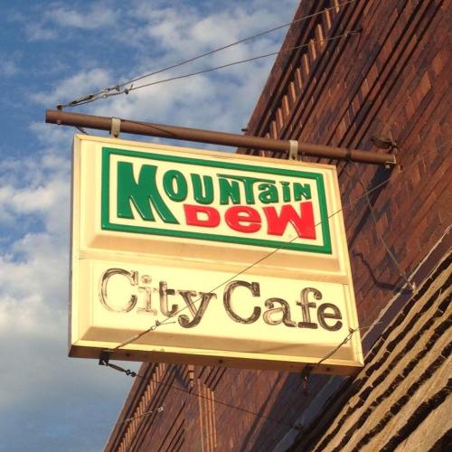 Sign for "Mountain Dew City Cafe" hanging from a brick building against a blue sky.