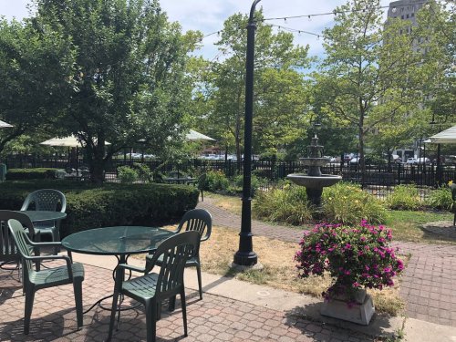 A peaceful outdoor area with green chairs, a table, a fountain, and blooming flowers surrounded by trees.