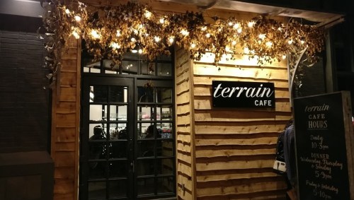 A cozy café entrance with wooden siding, warm lights, and a sign reading "terrain CAFE" above the door.