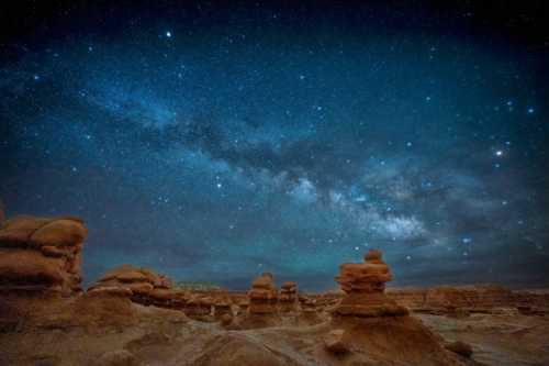 A starry night sky filled with the Milky Way over rocky formations in a desert landscape.