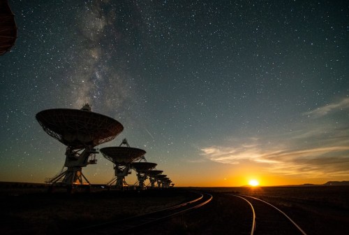 A row of radio telescopes under a starry sky, with the Milky Way visible and a sunset on the horizon.