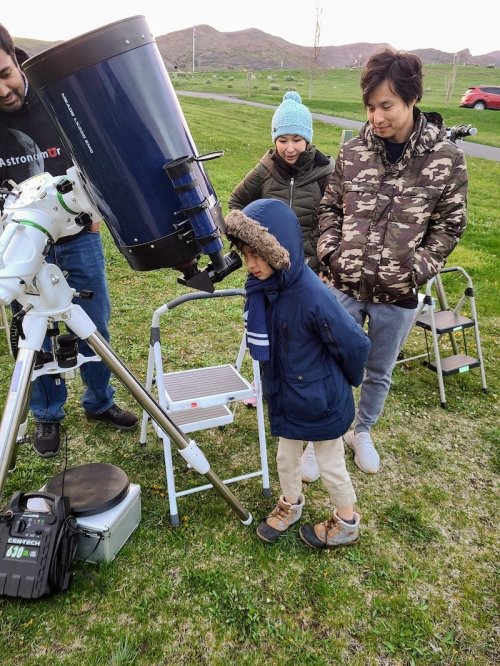 A child looks through a telescope while adults assist, set against a grassy field and distant hills.
