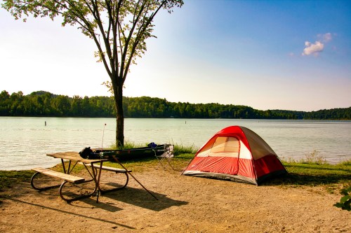 A red tent by a calm lake, with a picnic table and a kayak nearby, surrounded by trees under a clear blue sky.