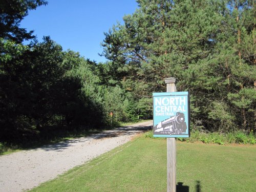Sign for North Central State Trail at a gravel path surrounded by trees under a clear blue sky.