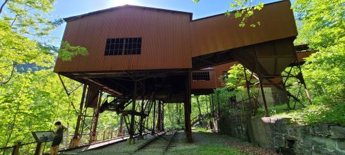 Abandoned industrial building surrounded by lush greenery and a gravel path, with a person observing nearby.