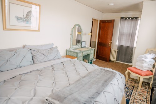 Cozy bedroom featuring a gray quilted bed, vintage dresser, and natural light from a window. Neatly stacked towels nearby.