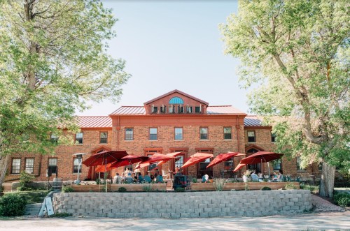 A brick building with a red roof, featuring outdoor seating under red umbrellas, surrounded by green trees.